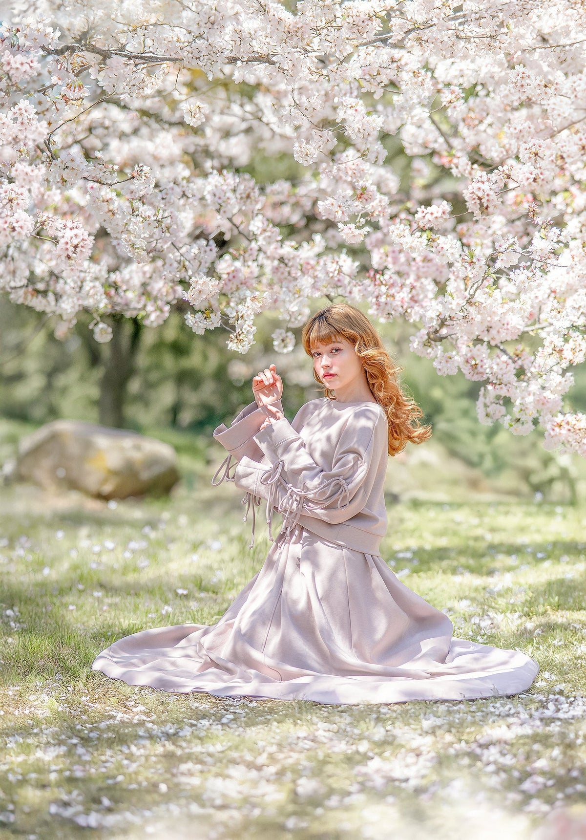 Reverie set in antique beige: a woman kneeling in the grass under a cherry blossom tree, bow-sleeve sweater and full skirt spreading around her.
