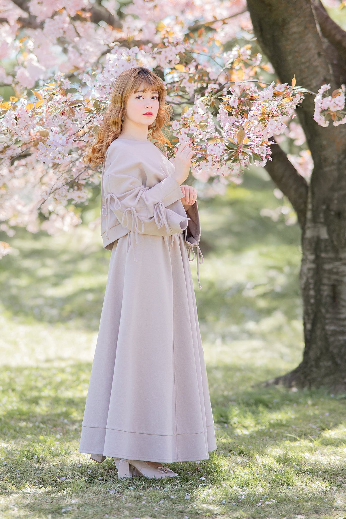 Reverie set in antique beige under a cherry blossom tree: bow-sleeve sweater with a long flowing skirt.