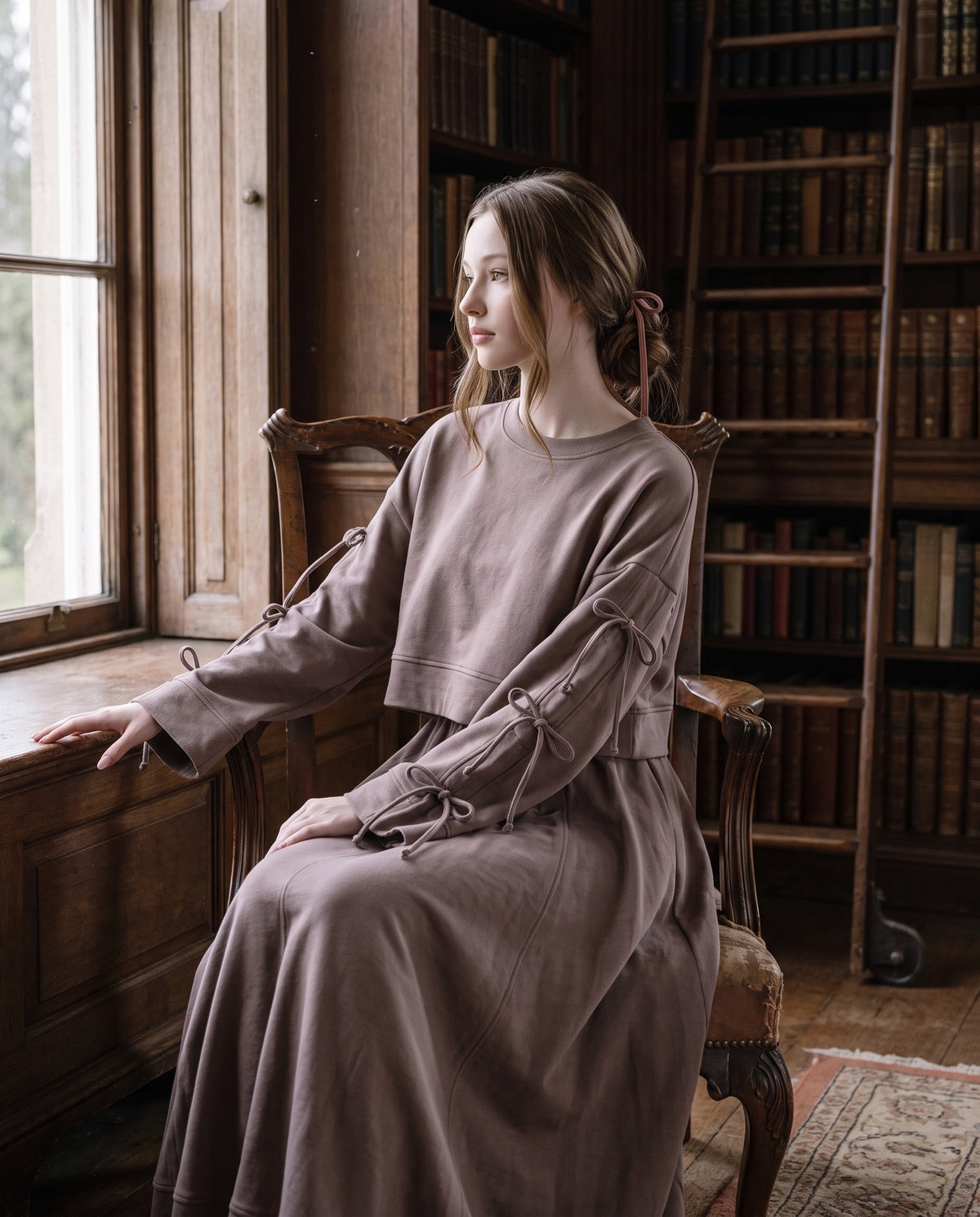 Young woman in the taupe Dustyrosa Reverie set, a cropped sweater with three bows on each sleeve and a long skirt, seated in a carved wooden armchair by a library window.