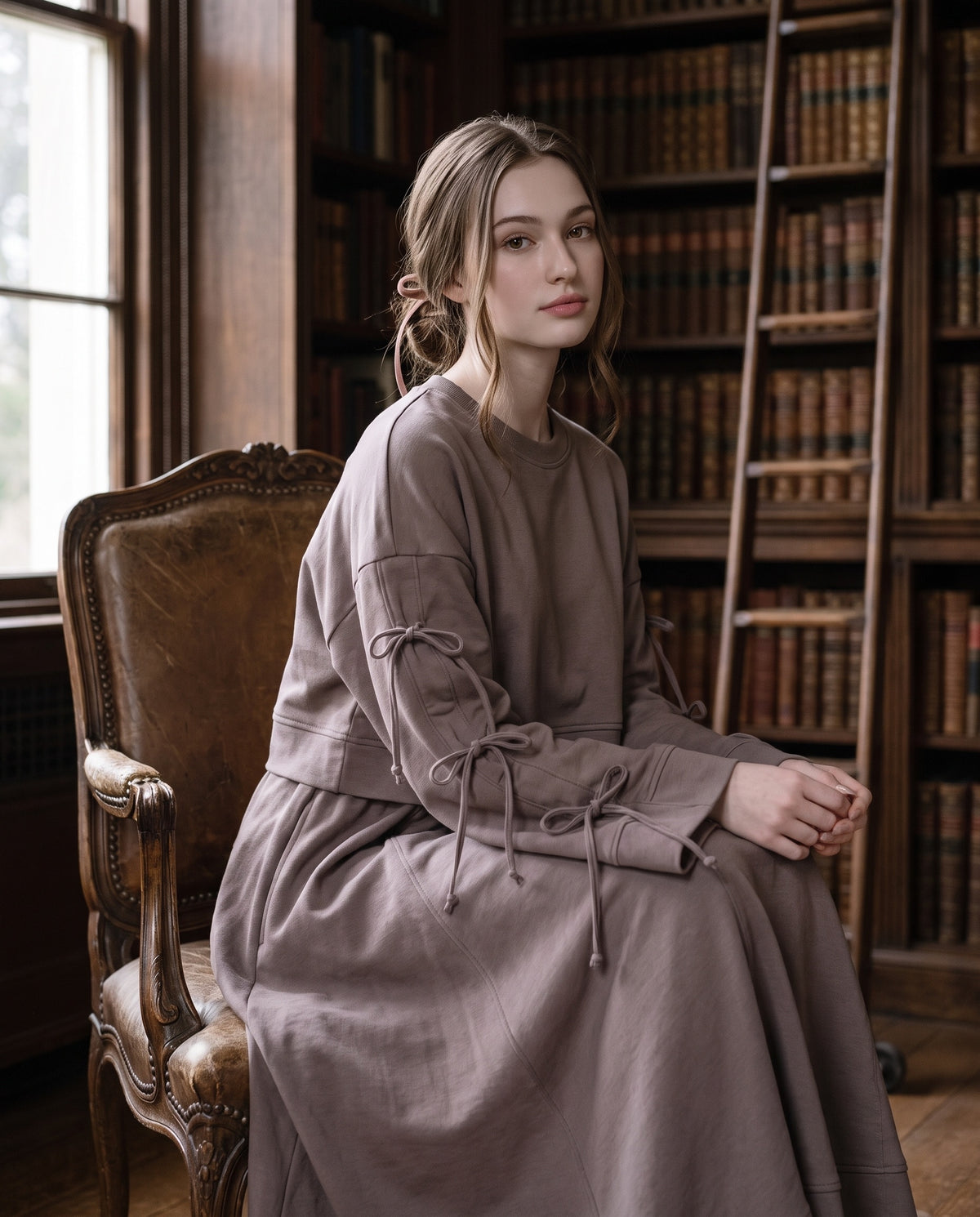Portrait of a woman in the taupe Dustyrosa Reverie set, seated on a leather armchair in front of tall bookshelves and a library ladder.