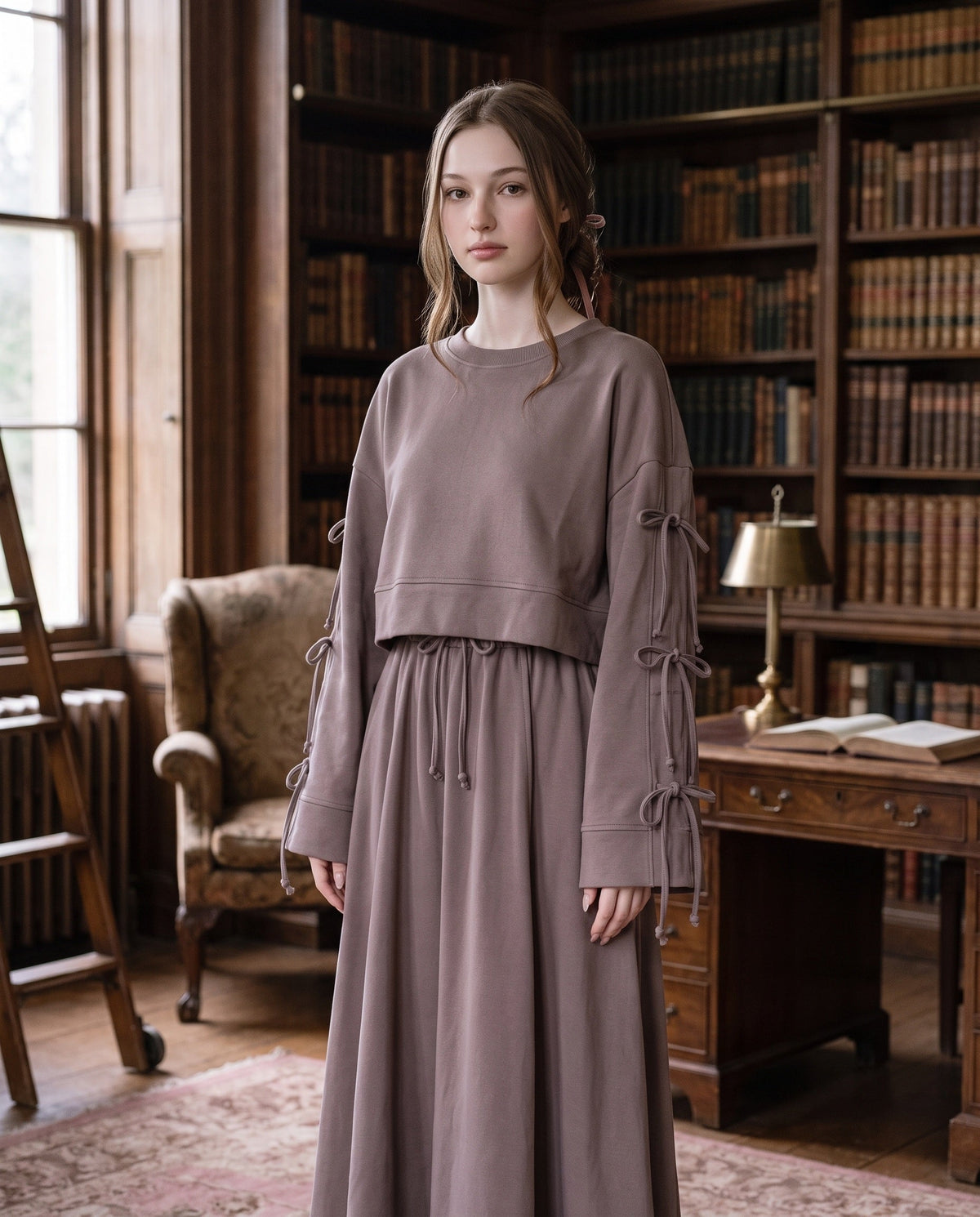 Woman standing in an old wood-panelled library wearing the taupe Dustyrosa Reverie set: cropped sweater with bows along the sleeves and a flowing long skirt with drawstring.